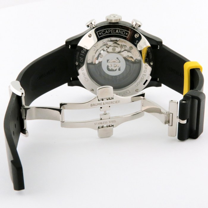 Back view of a Baume & Mercier wristwatch showing the transparent caseback and visible movement, with a black rubber strap and deployment clasp